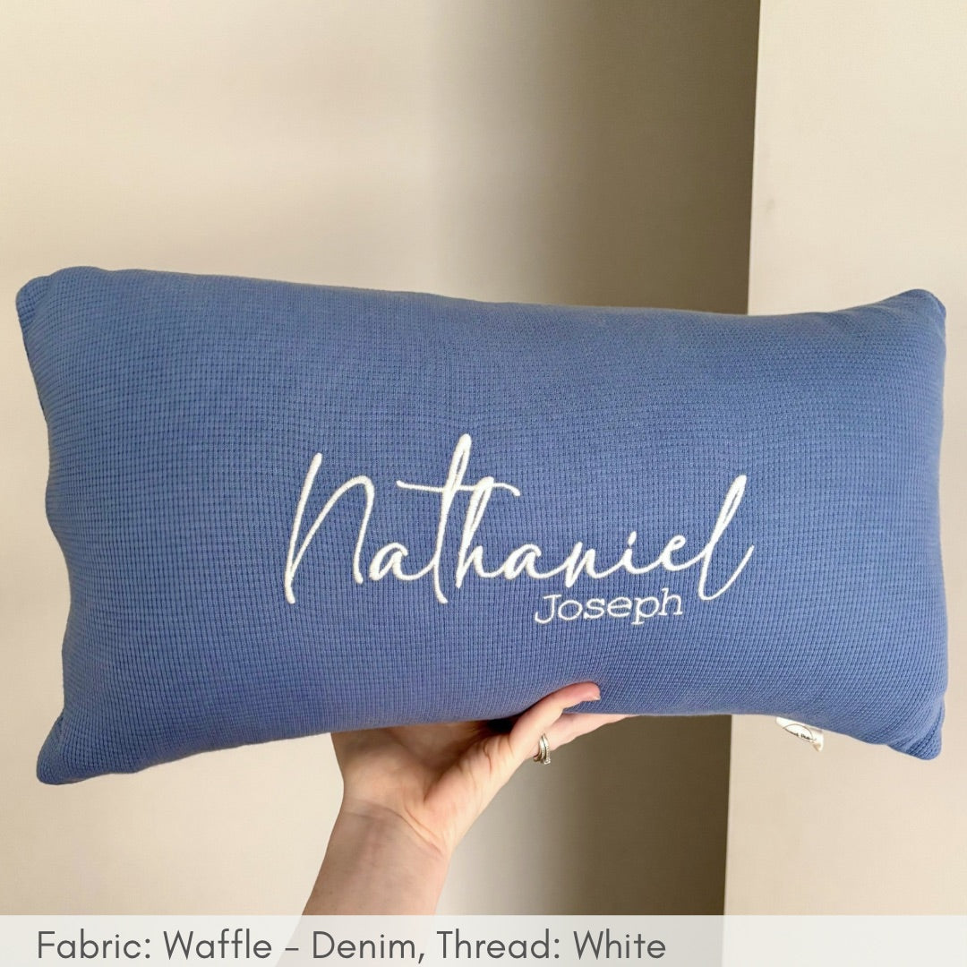 Blue pillow with 'Nathaniel Joseph' embroidered on it, held against a neutral background