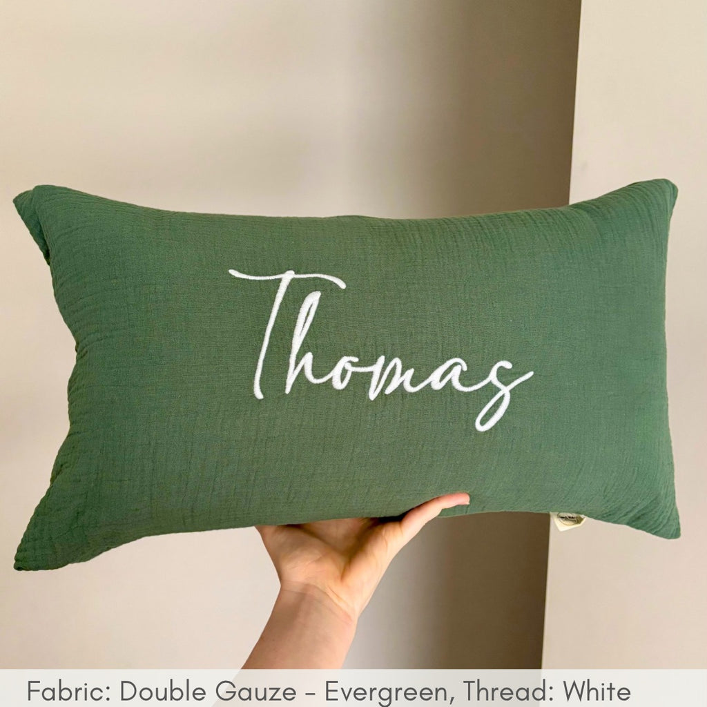 Green pillow with 'Thomas' printed on it held by a hand against a neutral background