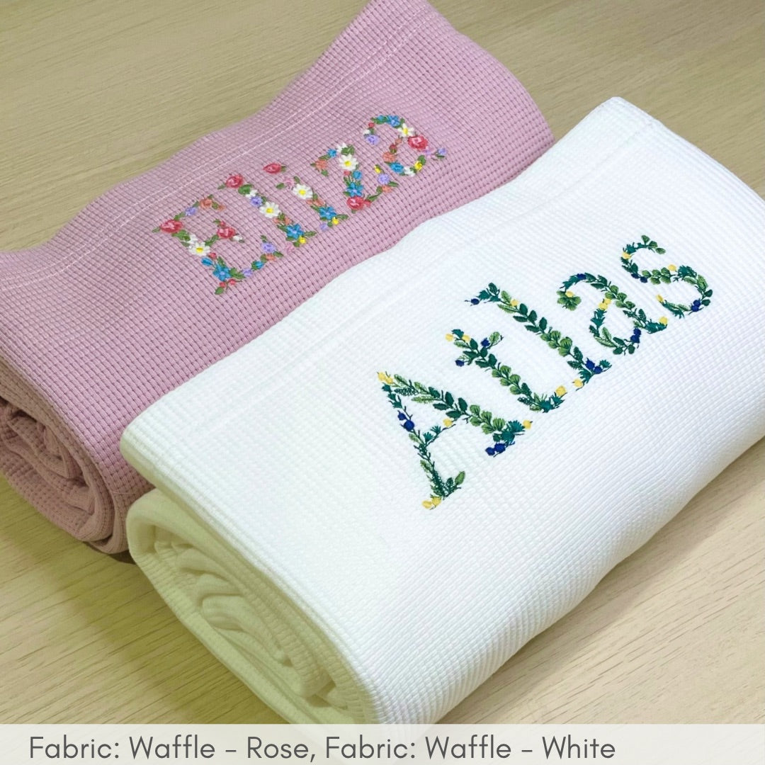 Two folded baby blankets with embroidered names on a wooden surface