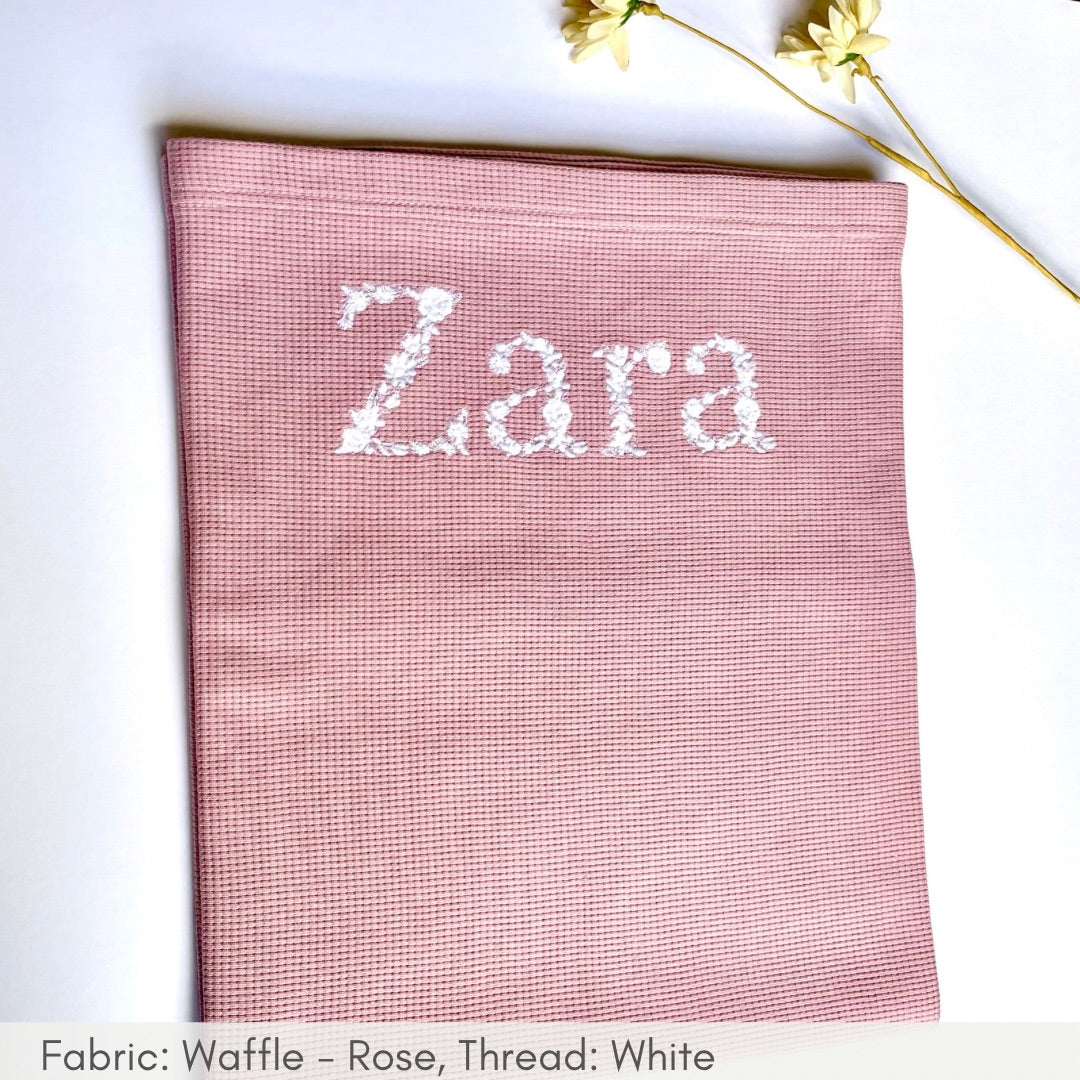 Pink fabric with 'Zara' embroidered on it, placed on a white surface with gold flowers.